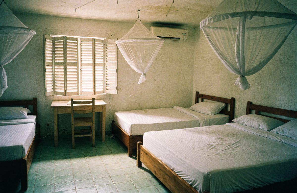 A large room with two beds under white mosquito nets, a desk by the window and a green tiled floor.