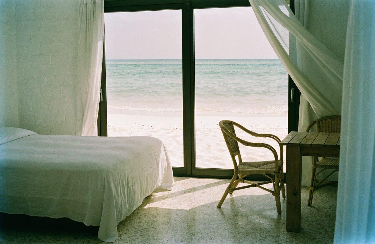 Sheer curtains drawn back from tall glass doors that open onto the beach and the sea.