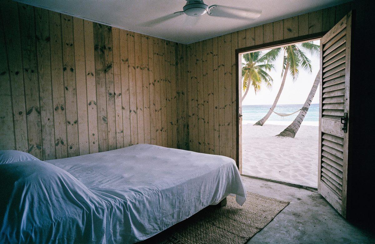 Leaning palms over a white sand beach, with the thatched roof of the house at the edge of the trees.